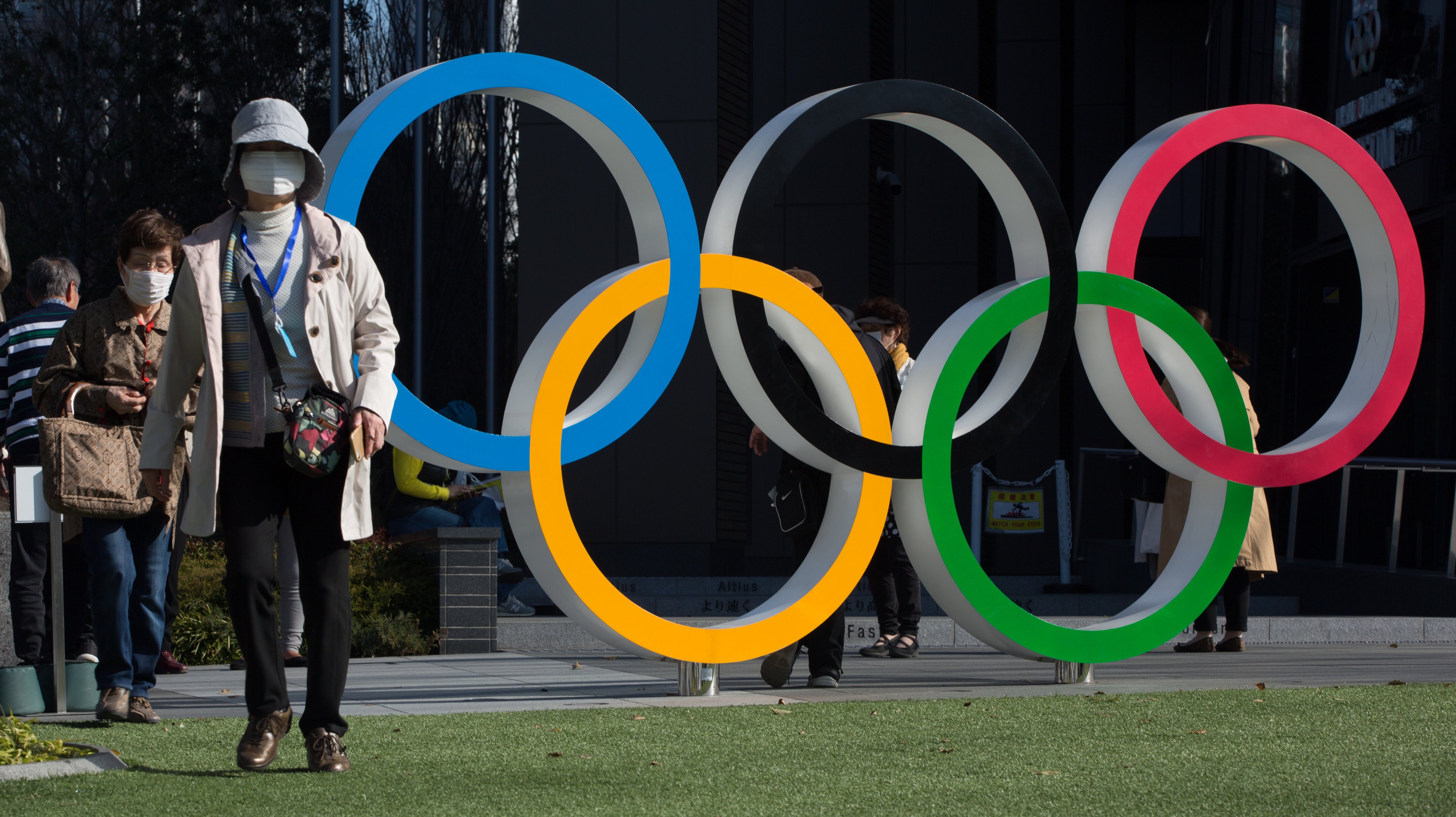 women with surgical masks walk next to the olympic rings in