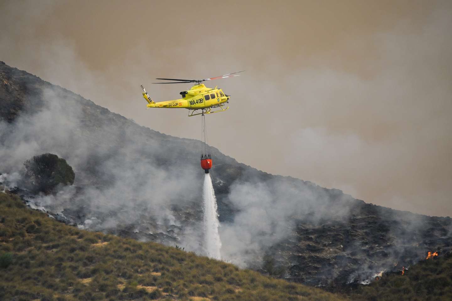 epa13102080 A fire service helicopter drops water onto one of the hotspots of the wildfire that broke out in Los Gallardos, Almeria, Spain, 10 July 2026. At least 12 people have died and 19 others are missing in a forest fire that broke out in evening of 09 July near Almeria in Andalusia, according to Andalusia's regional president Juanma Moreno.  EPA/CARLOS BARBA
