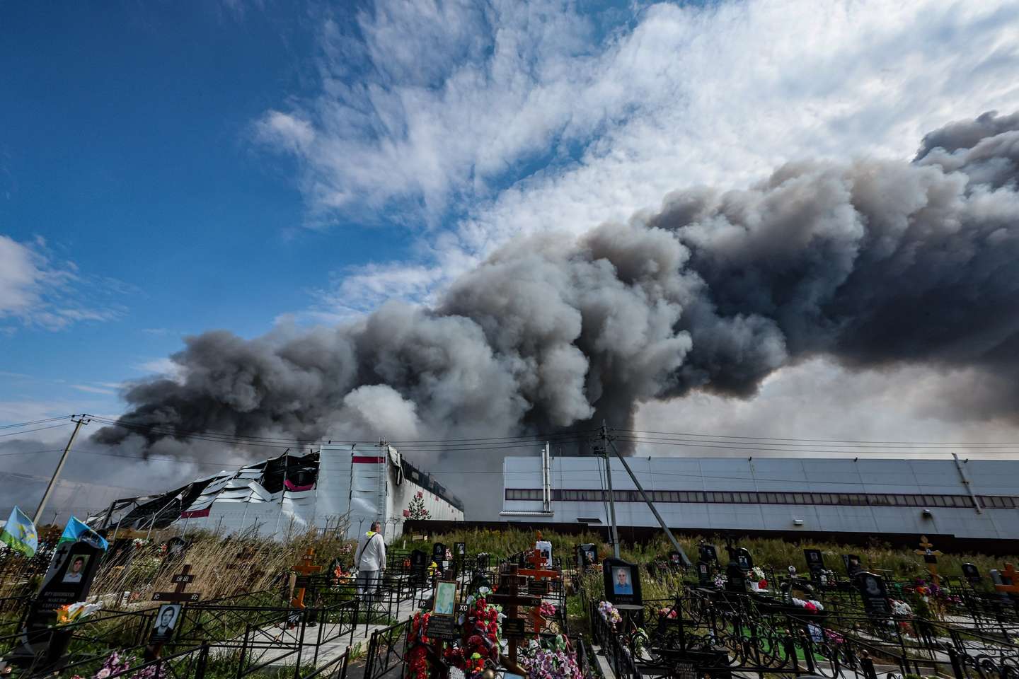 epa13173579 A person (C-L) stands among graves at a cemetery as smoke rises above a Wildberries facility (back) following a reported Ukrainian drone strike at the Koledino Industrial Park near Podolsk, Moscow Region, Russia, 16 August 2026. Moscow Region Governor Andrei Vorobyov reported that an overnight drone attack ignited a major fire at a warehouse belonging to Russia's largest online retailer Wildberries, adding that no employees were injured at the site.  EPA/STRINGER