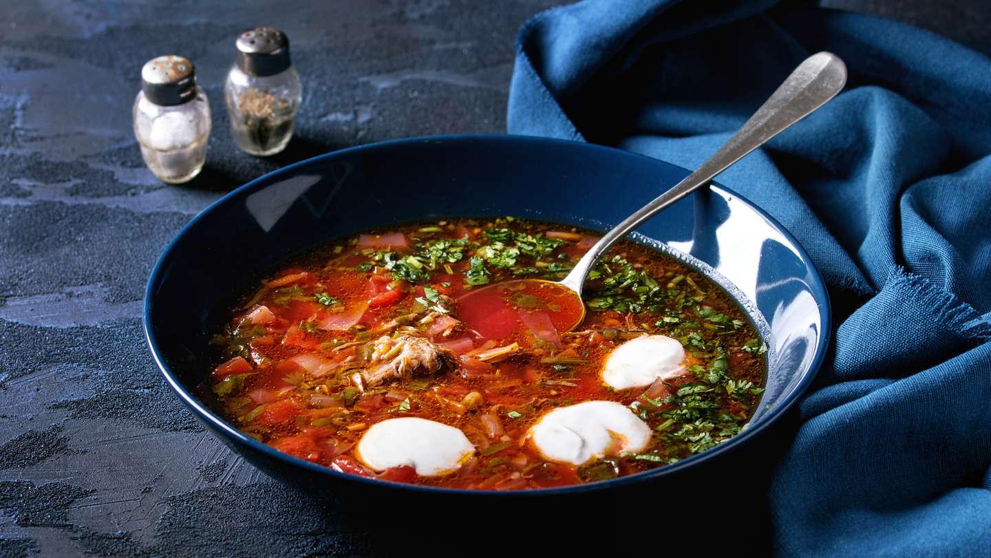 Plate of traditional beetroot borscht soup with sour cream and fresh coriander served with garlic bread buns pampushka with blue textile over dark blue texture background. Top view, space
