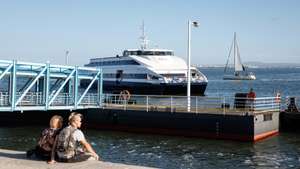 Um casal de turistas observa a chegada de um navio de passageiros, no Terreiro do Paço, em Lisboa, 10 de outubro de 2024. Cacilheiro. Catamarã. Transportes. Navio. Rio Tejo. Turismo. ANTÓNIO PEDRO SANTOS/LUSA