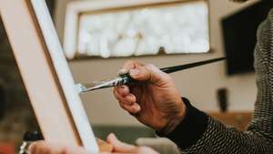 A man wearing glasses concentrates as he paints with oils onto a canvas on an easel