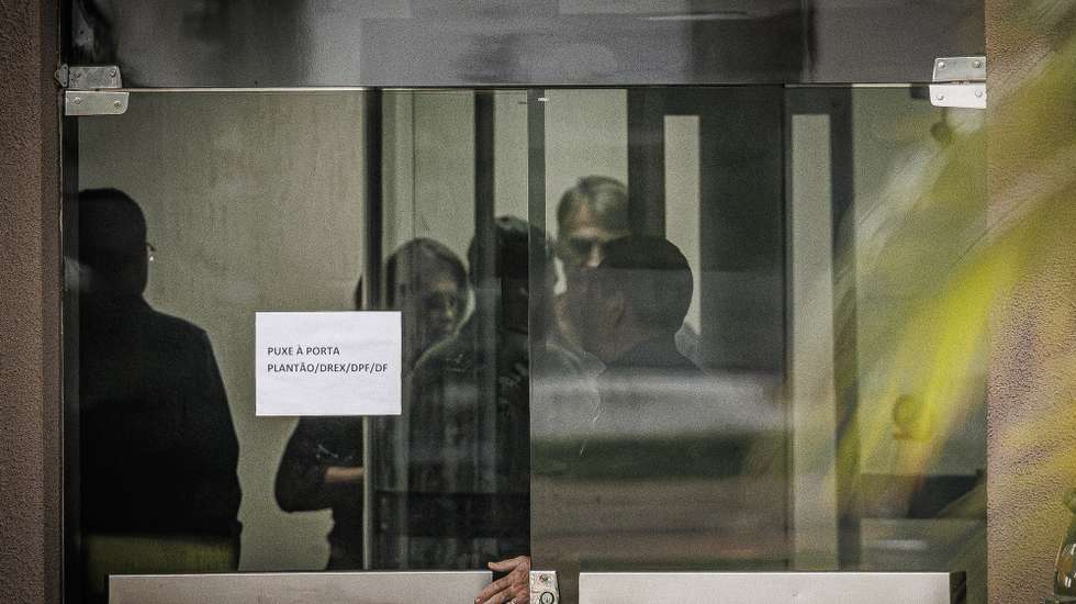 epaselect epa12544927 Former Brazilian President Jair Bolsonaro (rear-R) stands behind a glass door at Federal Police headquarters in Brasilia, Brazil, 23 November 2025. At a detention review hearing, Bolsonaro said a medication-induced episode of paranoia and hallucinations led him to tamper with and damage his electronic ankle monitor with a soldering iron because he believed it was a listening device.  EPA/Isaac Fontana
