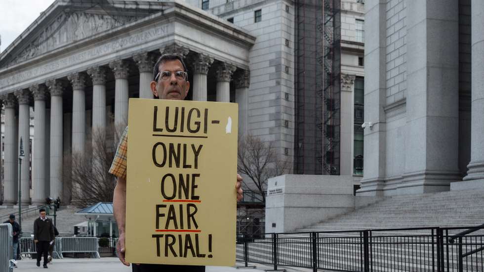 epa12865080 A supporter of Luigi Mangione holds a sign during Mangione's appearance in federal court in New York, New York, USA, 01 April 2026. A federal judge in New York pushed back Luigi Mangione's federal trial from September to October, giving an additional month's separation between his state and federal trials, which is set for June. Mangione is charged with second-degree murder and related counts in the 2024 killing of UnitedHealthcare CEO Brian Thompson.  EPA/OLGA FEDOROVA