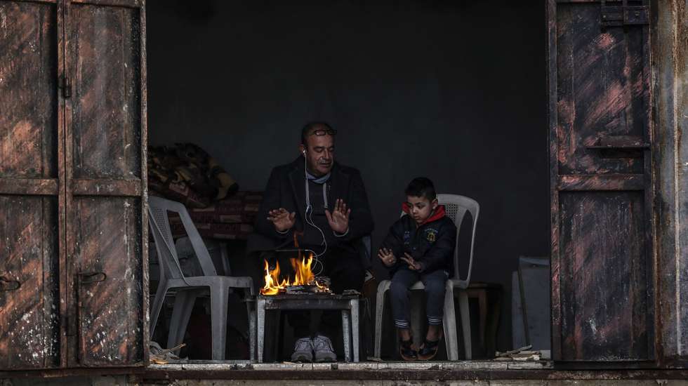 epa09707334 A Palestinian refugee and his son warm themselves by a fire in their family house during cold weather in the north of Al Shateaa Refugee Camp, Gaza Strip, 24 January 2022 (issued 25 January 2022). Most Palestinians in the Gaza Strip use batteries, genarators or candles to light their homes. Residents of Gaza, home to 1.8 million people, experience some 16 electricity outages per day. According to reports, the Gaza Power Generating Company plant usually operates only eight hours a day.  EPA/MOHAMMED SABER