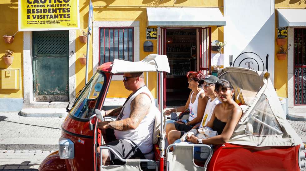 Um tuk-tuk transportando turistas passa por edifício com um cartaz com as palavras "Trânsito Caótico", da Associação QSintra -- Em Defesa de um Sítio Único, em Sintra, 16 de agosto de 2024. A Associação QSintra - Em Defesa de um Sítio Único - lançou uma iniciativa com faixas nas janelas e varandas e cartazes nas montras de lojas, restaurantes e cafés, exigindo à Câmara Municipal de Sintra medidas contra o "turismo de massas e caos no trânsito", com o objetivo de que a cidade não se transforme "num mero parque de diversões congestionado", pondo em causa o seu caráter de "sítio único". (ACOMPANHA TEXTO DA LUSA DO DIA 20 DE AGOSTO DE 2024). ANTÓNIO PEDRO SANTOS/LUSA
