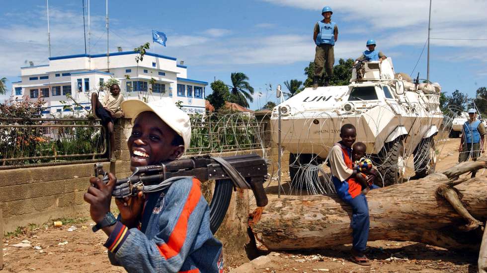 Child soldiers laugh outside the UN compound, 15 May 2003 in Bunia, Democratic Republic of Congo (DRC) as Un soldiers stand guard. The northeastern town of Bunia has been a theater of violent clashes between the Hema and the Lendu militias. MONUC (UN Mission to Congo) compound has been the main assembling point for the displaced from the region.  AFP PHOTO/MARCO L ONGARI             AFP PHOTO/MARCO L ONGARI