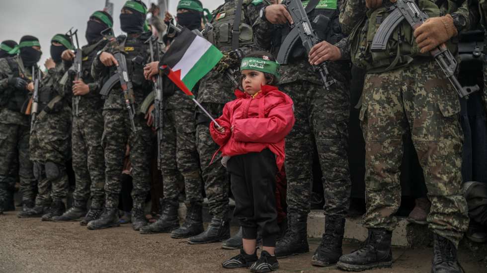 epa11915747 A Palestinian child holding a Palestinian flag stands near armed fighters from Hamas' Izz ad-Din al-Qassam Brigades during the handover of three Israeli hostages to Red Cross representatives in Al Nuseirat refugee camp, central Gaza Strip, 22 February 2025. Six Israeli hostages were released on 22 February -- three in Nuseirat, two in Rafah and one in Gaza City -- after Hamas handed them over to the Red Cross in Gaza as part of the first phase of the Israel-Hamas ceasefire deal.  EPA/MOHAMMED SABER