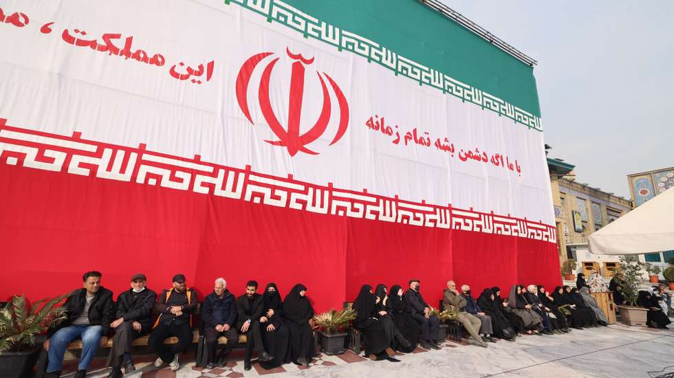 epa12819685 People and relatives of late Iranian supreme leader's top adviser Ali Shamkhani sit under a huge Iran national flag during a funeral ceremony at the Saleh shrine in Tehran, Iran,14 March 2026. A joint Israeli and US military operation continues to target multiple locations across Iran since the early hours of 28 February 2026.  EPA/ABEDIN TAHERKENAREH