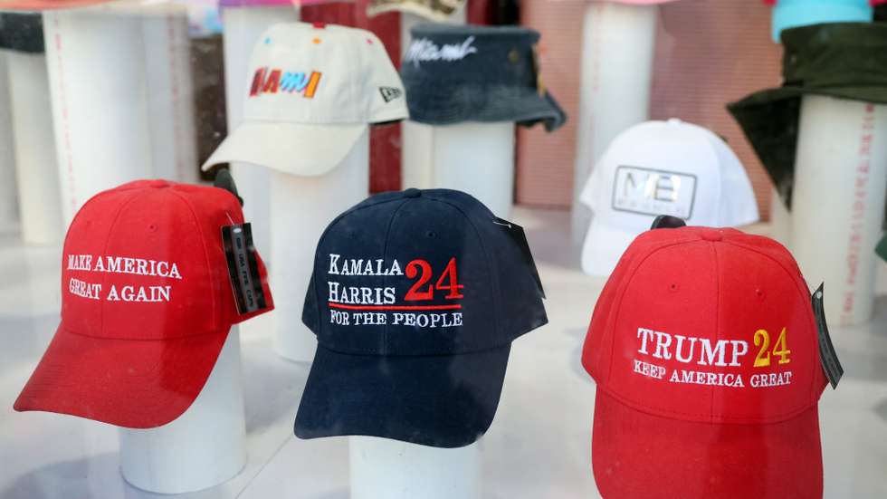 Uma montra de uma loja de roupa expõe os bonés oficiais das campanhas de Kamala Harris e Donald Trump, perto de uma mesa onde decorre o voto antecipado para as eleições americanas, Miami, 25 de outubro de 2024. É sob um intenso sol caribenho e desafiando temperaturas superiores a 30.ºC que dezenas de eleitores se deslocam este final de semana ao edifício-sede do município de Miami, um dos dezenas de locais de voto antecipado no sul deste estado com perto de 22 milhões de habitantes. (ACOMPANHA TEXTO DE 26 DE OUTUBRO DE 2024. NUNO VEIGA/LUSA