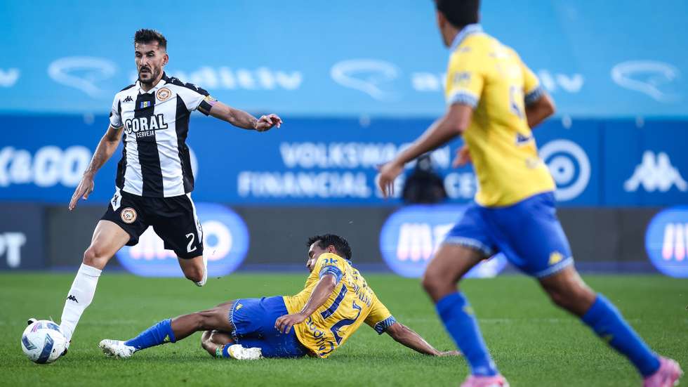 O jogador do Estoril Praia, João Carvalho (C), em ação contra o jogador do Nacional, João Aurélio (E), durante o jogo da Primeira Liga de Futebol, disputado no Estádio António Coimbra da Mota, no Estoril, Cascais, 26 de outubro de 2025. RODRIGO ANTUNES/LUSA