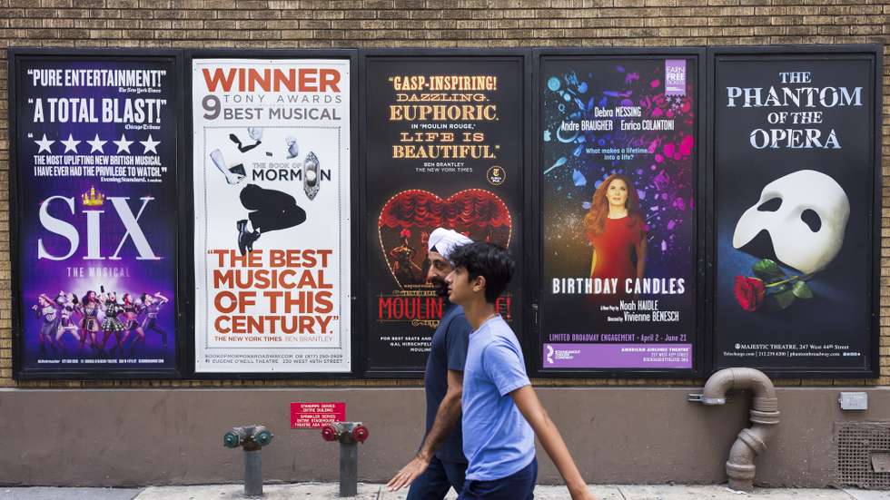 epa09380662 People walk past posters for musicals in the Broadway theater district near Times Square in New York, New York, USA, 30 July 2021. The owners and operators of the city's theaters and performance venues announced on 30 July 2021 that when plays, musicals and other in-person shows restart next month, audience members will be required to show proof of COVID-19 vaccination and wear face masks.  EPA/JUSTIN LANE