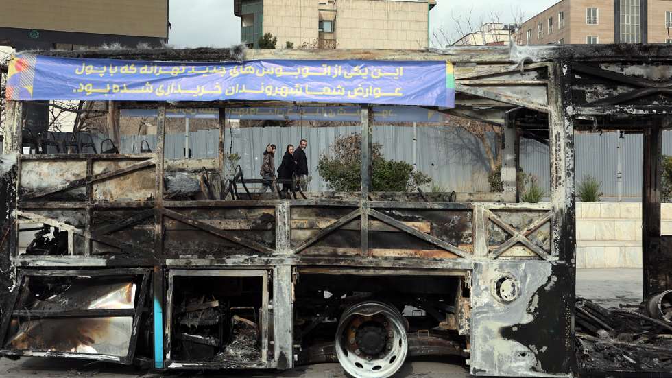 epa12650627 Iranians walk near the wreckage of a burnt-out public bus in Tehran, Iran, 15 January 2026. The country remains under a near-total nationwide internet blackout that began on 08 January, amid an intensifying wave of anti-government protests.  EPA/ABEDIN TAHERKENAREH