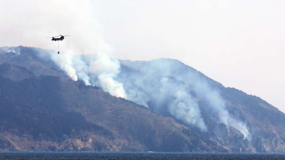 epa11929164 A military helicopter assists with firefighting efforts as smoke rises from the hills in Ofunato, Iwate prefecture, northeastern Japan, 28 February 2025. A body was found in Ayazato, Sanriku Town, Iwate Prefecture as the area destroyed by the fire is over 600 hectares. At least 84 buildings have been destroyed by the fire, and local firefighters are continuing firefighting efforts.  EPA/JIJI PRESS JAPAN OUT EDITORIAL USE ONLY