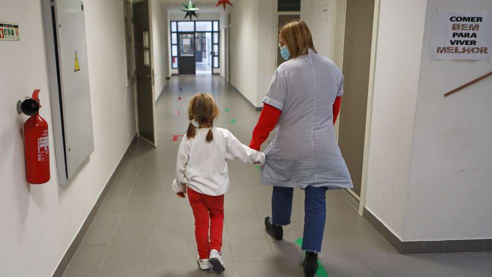 Uma educadora de infância, com uma criança, percorre um corredor da Escola Básica de São Vicente, que vai funcionar até 07 de janeiro de 2022, como escola de acolhimento, em Telheiras, Lisboa, 30 de dezembro de 2021. O Ministério da Educação voltou a pôr em funcionamento desde dia 27 de dezembro a rede de escolas de acolhimento para os filhos, ou outros dependentes, de trabalhadores essenciais. ANTÓNIO PEDRO SANTOS/LUSA