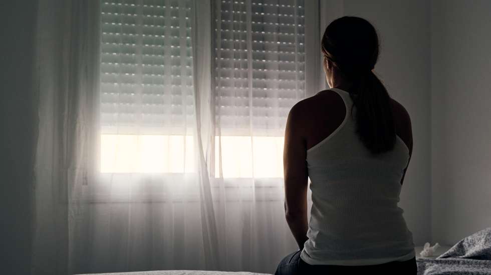 Rear view of an unrecognizable abused woman sitting on her bed looking out the window.
