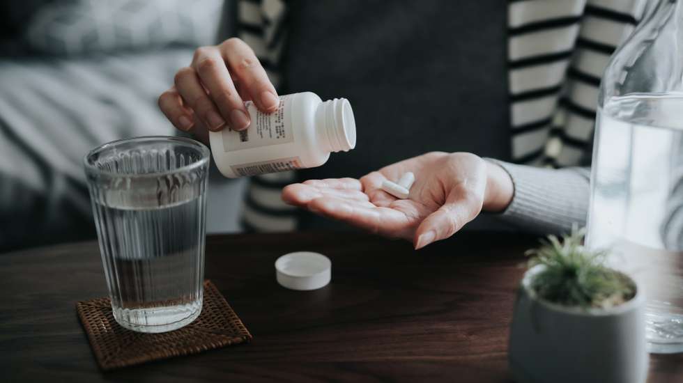 Close up of young Asian woman holding a pill bottle, pouring pills into palm of hand, with a glass of water on the side. Medicine, healthcare and people concept