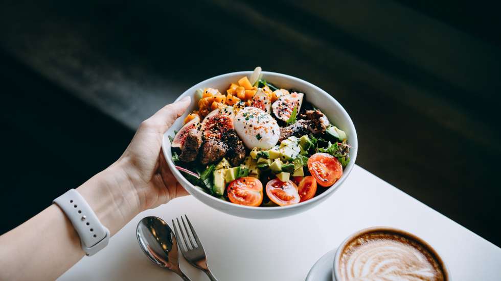 Close up of woman's hand holding a bowl of fresh beef cobb salad, serving on the dining table. Ready to enjoy her healthy and nutritious lunch with coffee. Maintaining a healthy and well-balanced diet. Healthy eating lifestyle