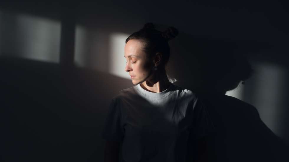 A lonely Young Sad Calm Girl Or Woman stands against a white wall, in a psychiatric clinic or at home, in the rays of sunlight, with her eyes closed. The Concept of Depression from Illness and Quarantine, Fatigue and Impotence. A copy of the space.