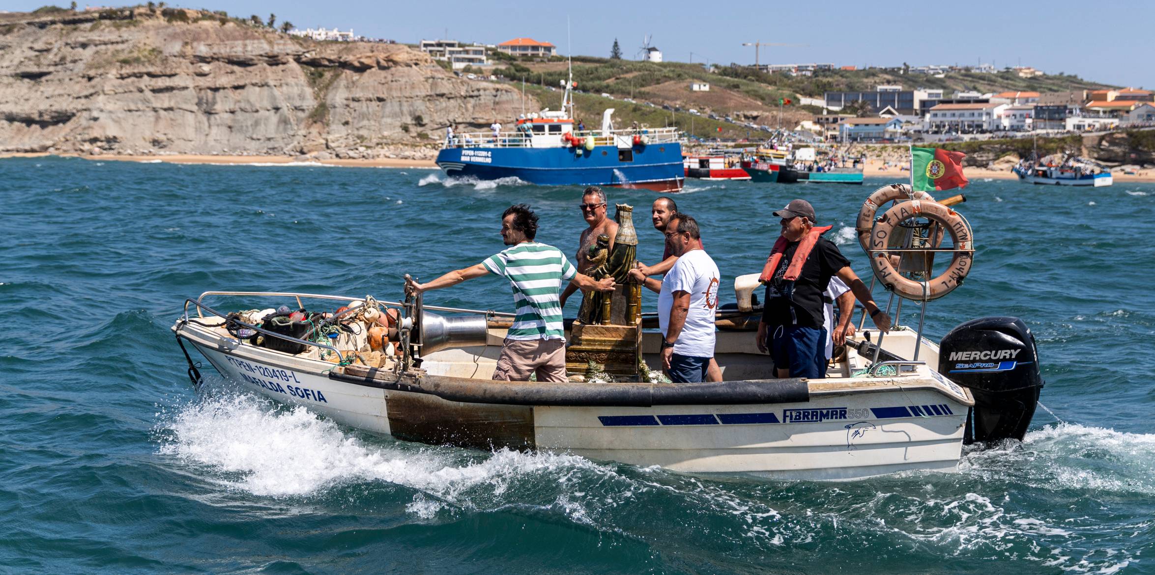 Em Porto Dinheiro: ao longe veem-se centenas de pessoas, enquanto uma lancha traz a estátua prometida. Os barcos apitam desenfreadamente e disparam foguetes à passagem da lancha