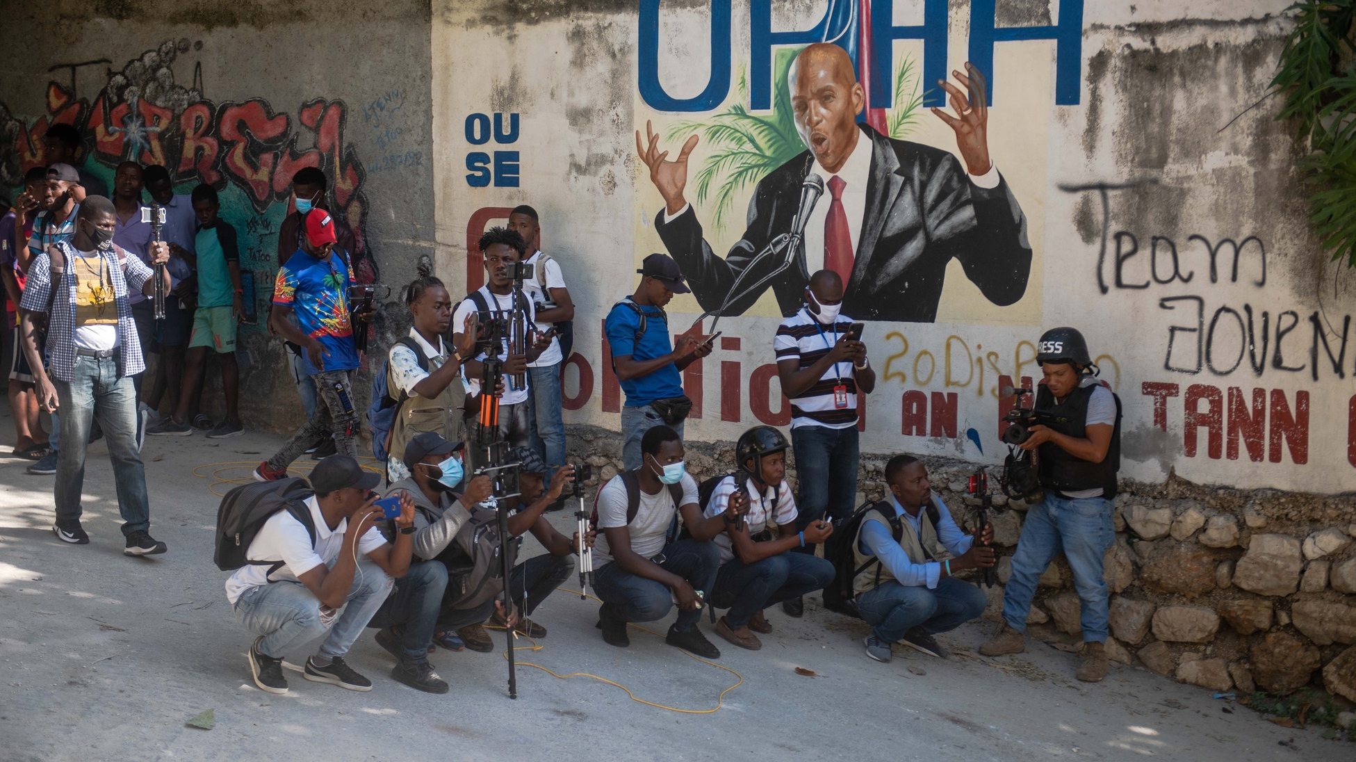 epa09329422 A group of journalists work next to a mural of the assassinated president Jovenel Moise, in Port-au-Prince, Haiti, 07 July 2021. The President of Haiti Jovenel Moise, was assassinated on 07 July by armed men who carried out an attack on his residence in the early morning in the Pelerin neighborhood of Port-au-Prince, said interim prime minister, Claude Joseph.  EPA/JEAN MARC HERVE ABELARD