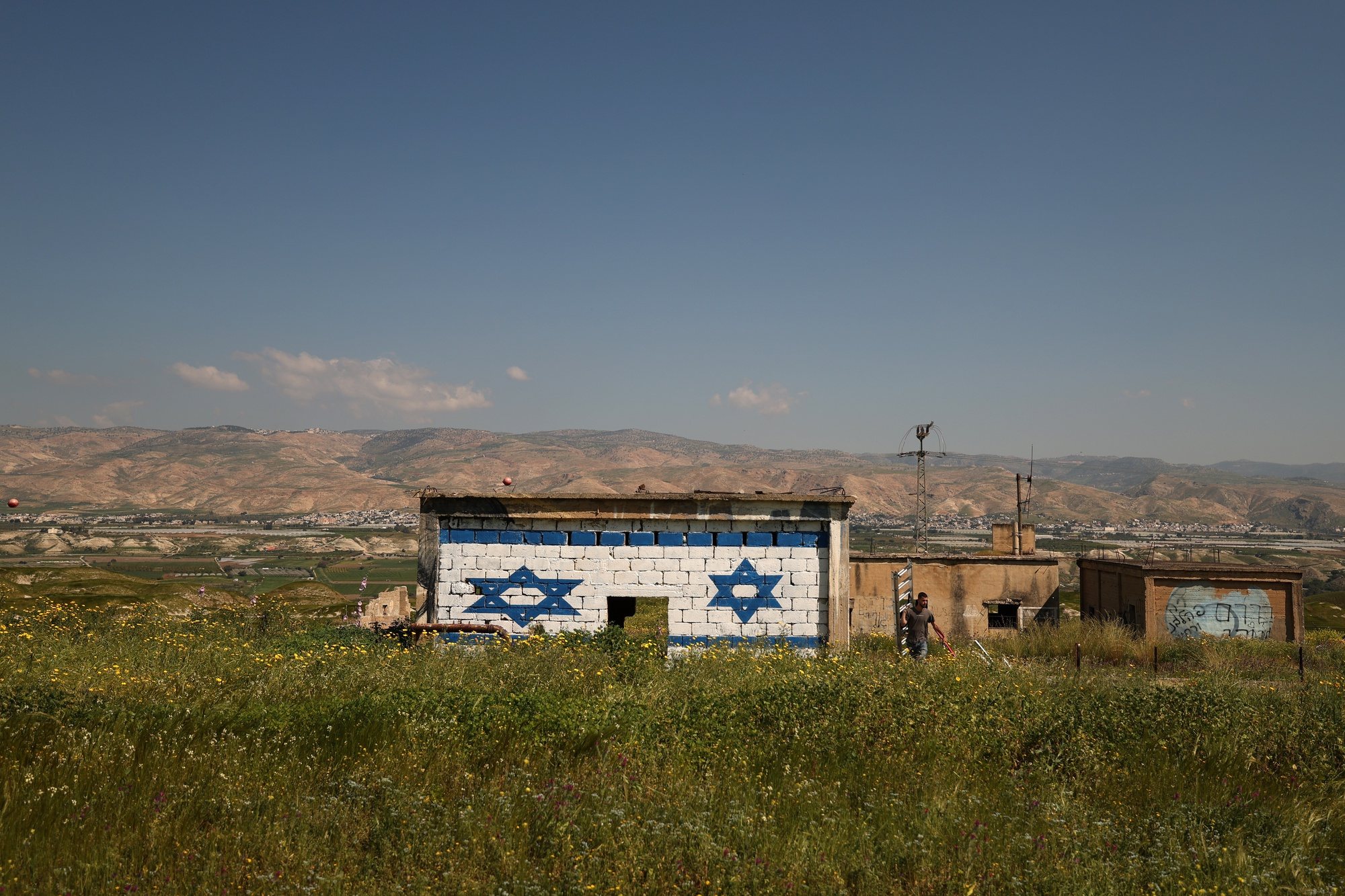 epa11214811 An Israeli right-wing activist walks past an abandoned West Bank house after painting it with an Israeli national flag, in the Jordan Valley, as the city of Ajlun, Jordan, is seen in the background, 11 March 2024.  EPA/ATEF SAFADI