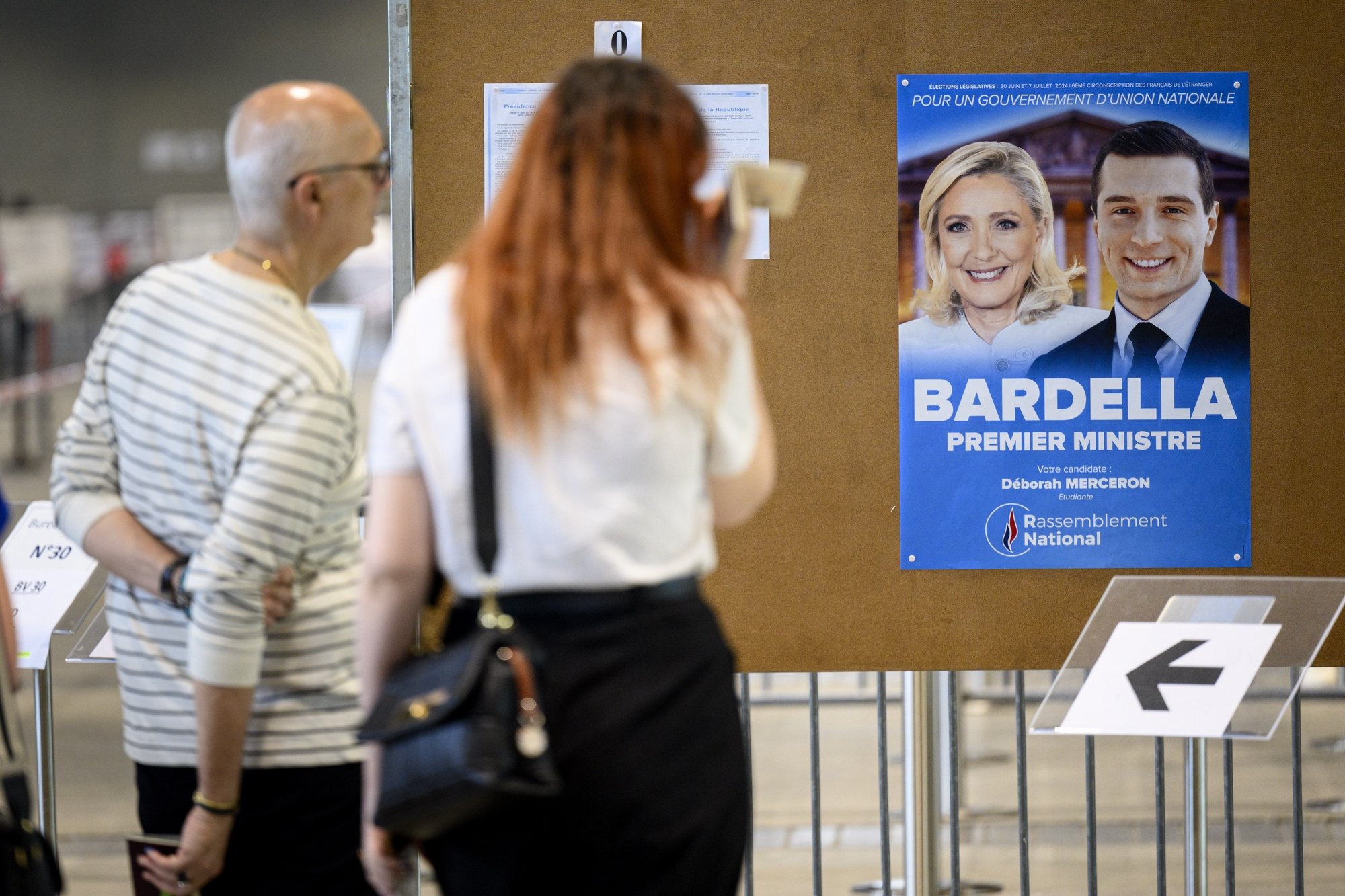 epa11447128 French people living in Switzerland look at an election poster showing French member of parliament Marine Le Pen (L) and French far right party National Rally (RN) leader Jordan Bardella (R) as they line up to cast their ballots during the first round of the French parliamentary elections, at a polling station in Lausanne, Switzerland, 30 June 2024. France on 30 June holds the first round of snap parliamentary elections called by President Emmanuel Macron, after dissolving the National Assembly on 09 June 2024.  EPA/LAURENT GILLIERON