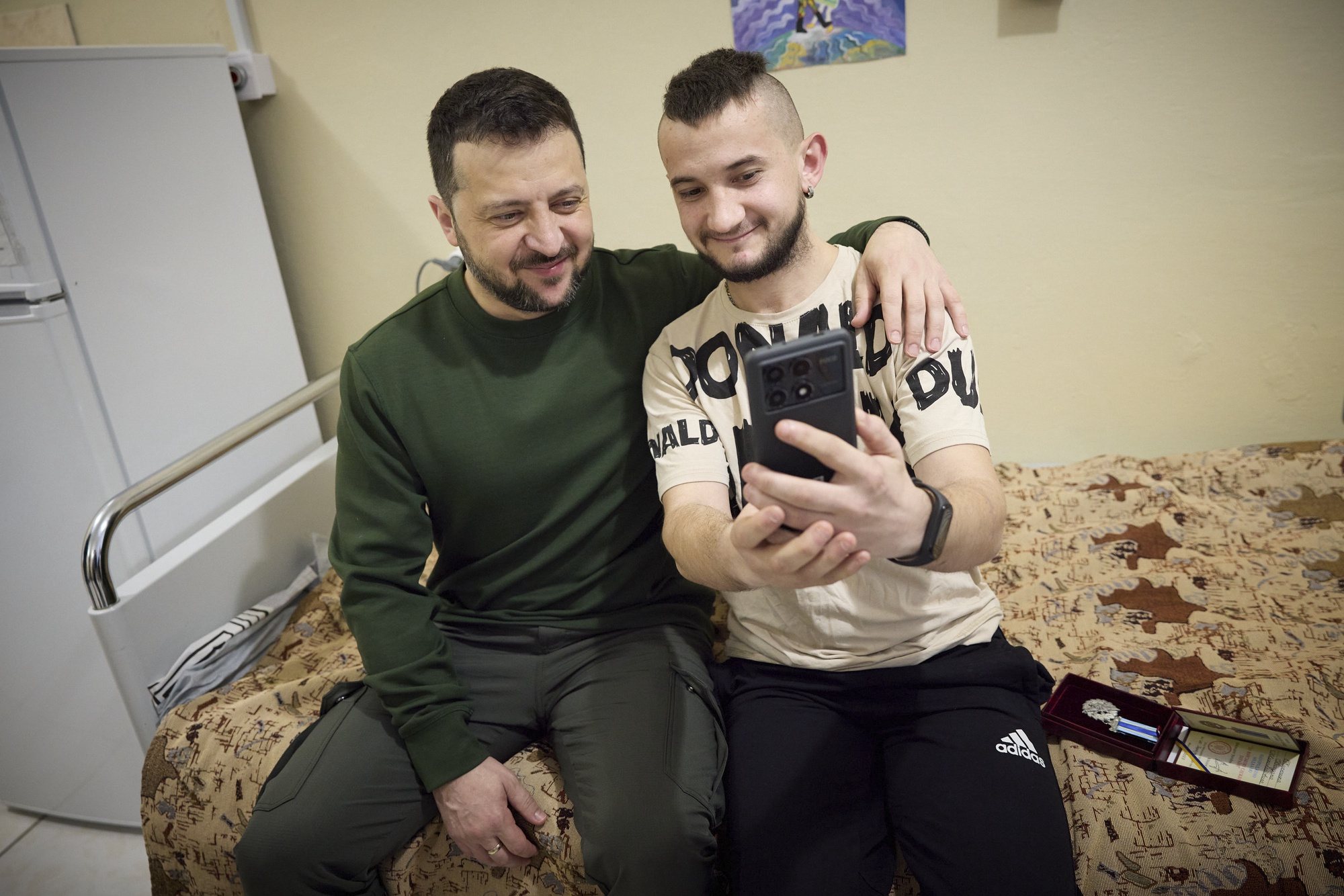 epa11247107 A handout picture made available by the Presidential Press Service shows Ukrainian President Volodymyr Zelensky (L) posing for a selfie with a wounded serviceman at a military hospital during a working visit to the Sumy region, Ukraine, 27 March 2024 amid the Russian invasion. Zelenskyy was briefed on the organization across three lines of defense and discussed the current needs for weapons and equipment. Russian troops entered Ukrainian territory on 24 February 2022, starting a conflict that has provoked destruction and a humanitarian crisis.  EPA/PRESIDENTIAL PRESS SERVICE HANDOUT   HANDOUT EDITORIAL USE ONLY/NO SALES HANDOUT EDITORIAL USE ONLY/NO SALES