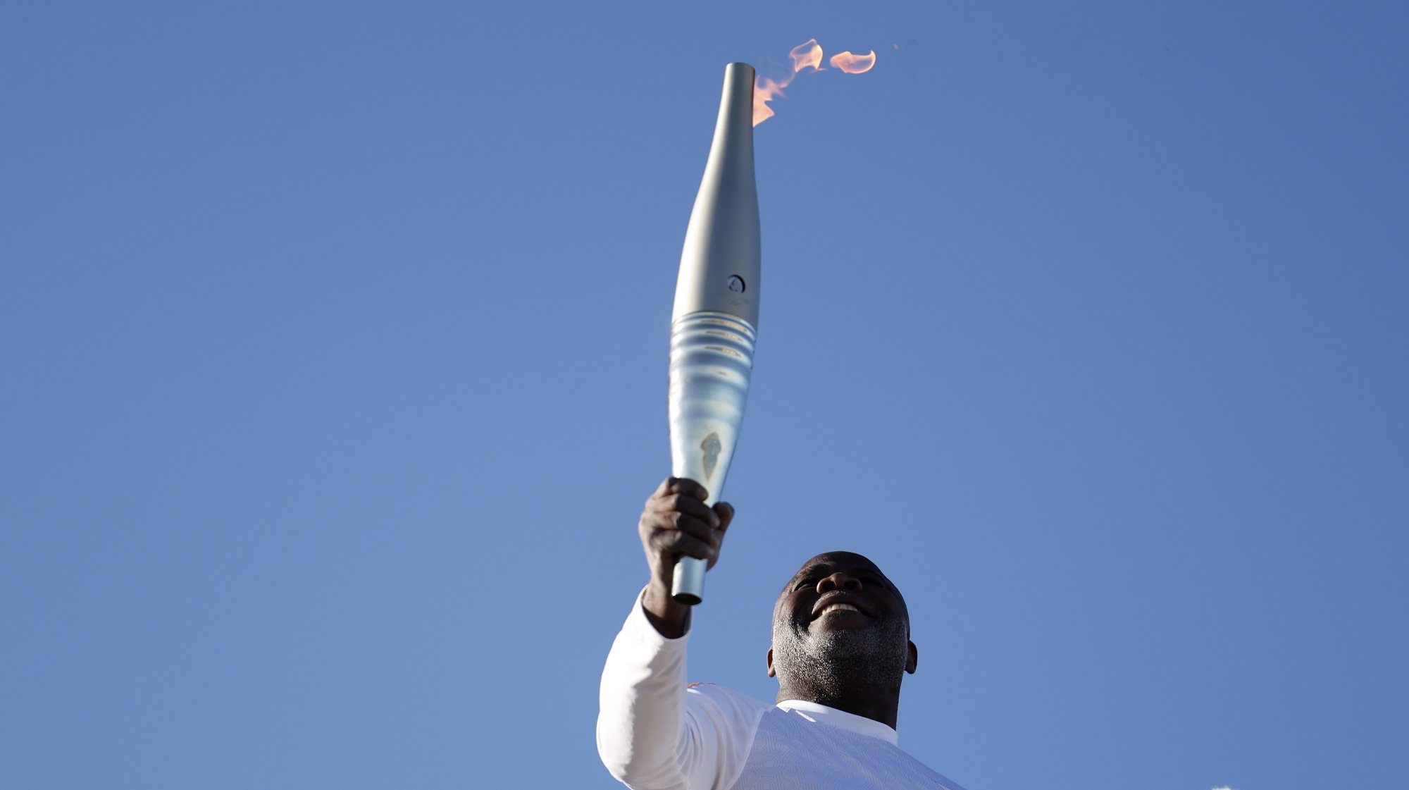 epa11328363 Former football player Basile Boli holds the Olympic Flame at Notre Dame De La Garde cathedral in Marseille, France, 09 May 2024, for the first leg of Olympic Torch relay. The Olympic Flame arrived in Marseille on 08 May following a 12-day journey from Piraeus, Greece, on board the Belem, a three-masted sailing ship built in 1896 and currently serving as a training ship under the French flag. The Paris 2024 Olympic Games will start on 26 July 2024.  EPA/GUILLAUME HORCAJUELO