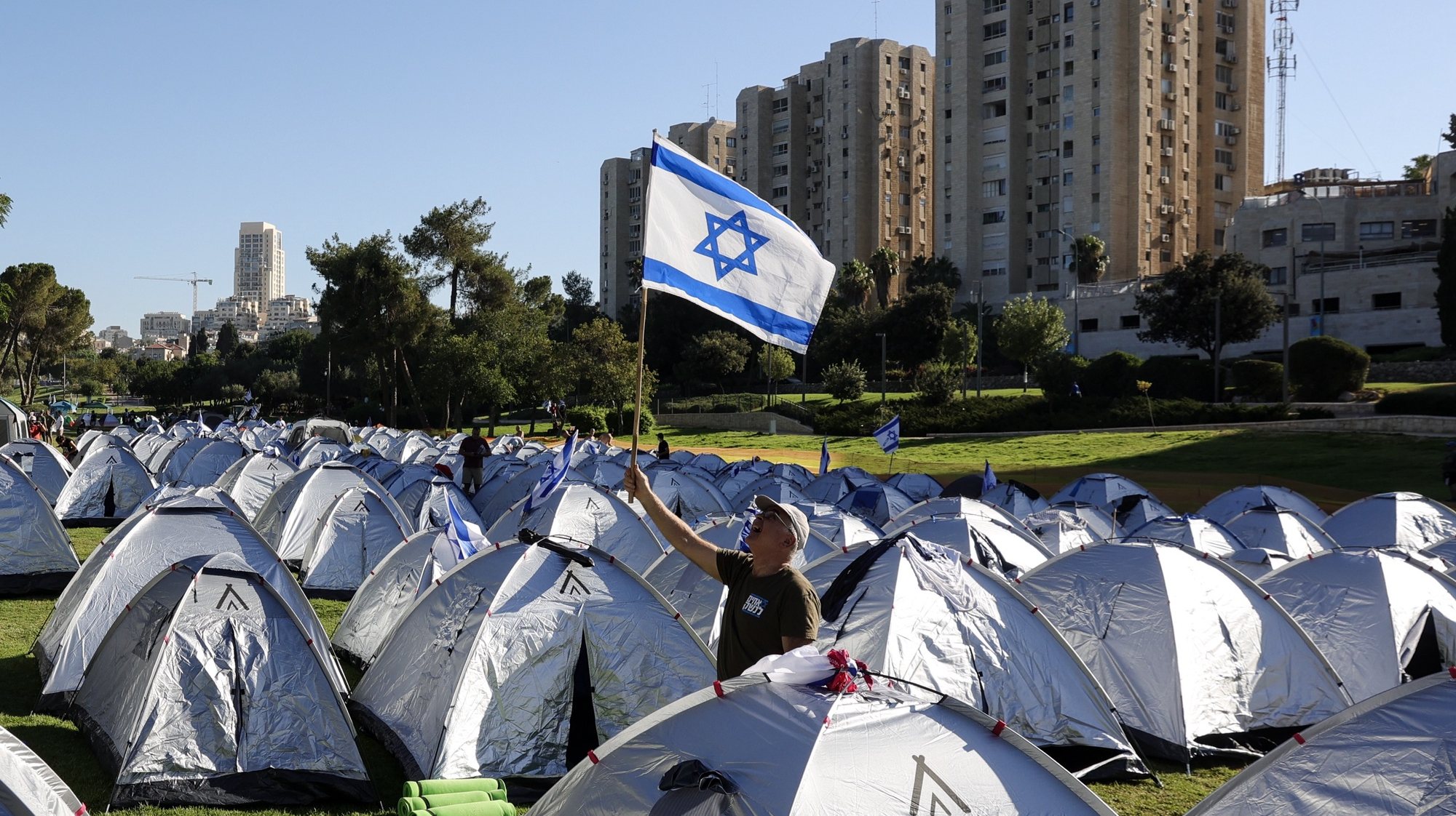 epa10763324 An anti-government raises a Israeli flag among tents after spending the night in a tent camp at Sacher Park, near the Israeli Knesset, following a four-day protest march to Jerusalem against the government&#039;s planned justice system reform, in Jerusalem, 23 July 2023. Mass protests continue across the country as the Israeli parliament, or Knesset, is set to hold a final vote on 23 and 24 July on the bill that would limit the Supreme Court&#039;s powers.  EPA/ABIR SULTAN