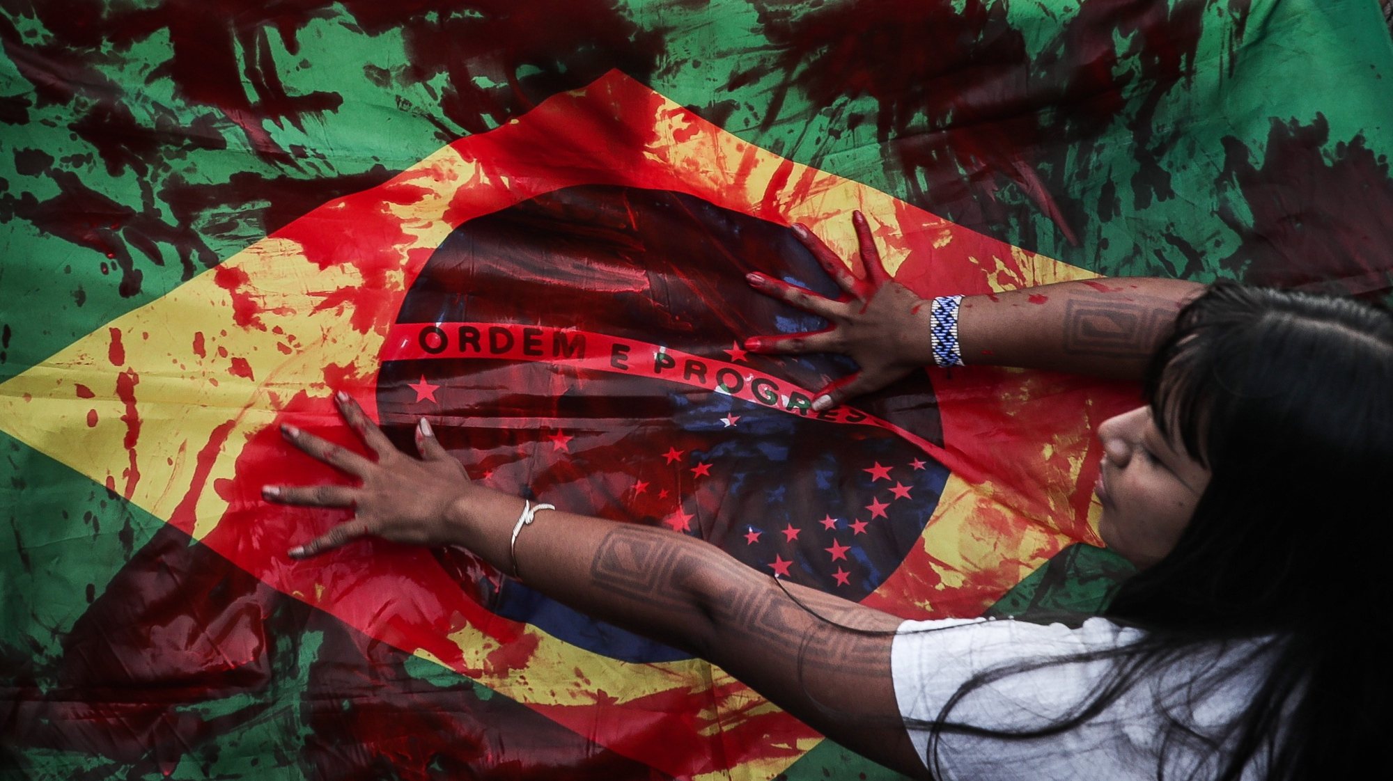epa10663928 A Brazil national flag is smeared in red paint as indigenous people protest against a bill that makes it difficult to demarcate land in Brazil, at the Ministries Esplanade, in Brasilia, Brazil, 30 May 2023. A group of indigenous people of the Guarani ethnic group blocked one of the main highways of Sao Paulo, the most populous of the Brazilian states, in protest against a bill that is being processed in Congress and that could make it difficult to demarcate the native lands.  EPA/ANDRE COELHO