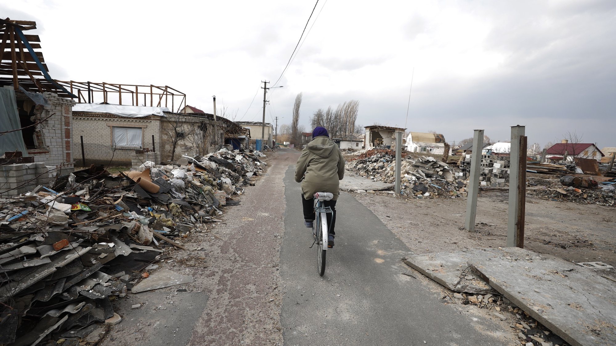 epa09857812 A woman rides a bicycle past damaged houses in the aftermath of a Russian airstrike in the village of Ulica Szkolna, Kyiv Oblast, 29 March 2022. Russian troops entered Ukraine on 24 February resulting in fighting and destruction in the country, and triggering a series of severe economic sanctions on Russia.  EPA/ATEF SAFADI