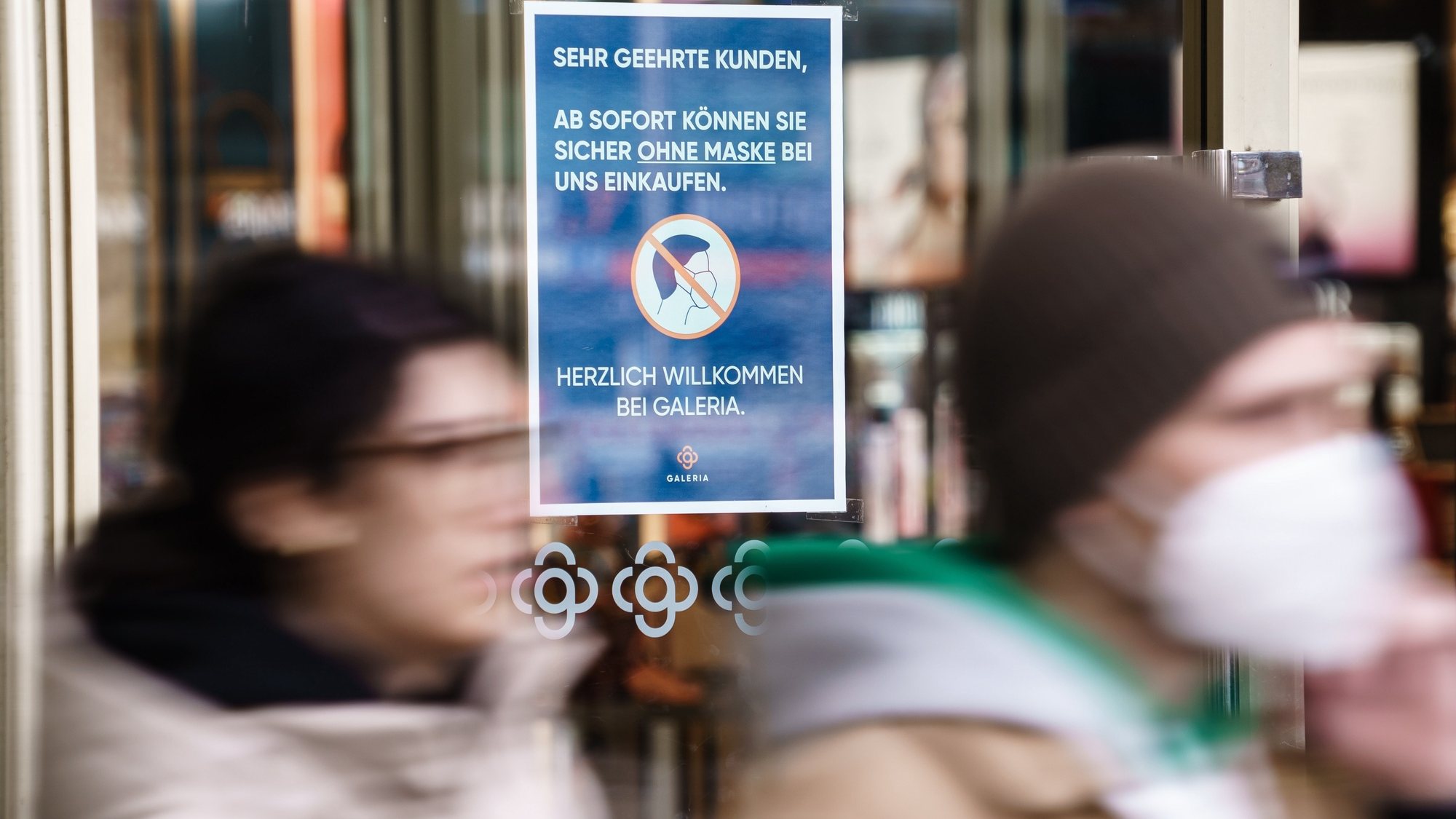 epa09864065 A person with a face mask on (R) and another person without, leave a Galeria department store, in front of a note reading in German language ’Dear customers, as of now you can safely shop with us without a mask - welcome at Galeria.’ in Berlin, Germany, 01 April 2022. Berlin drops most coronavirus related measures from 01 April 2022, among them the necessity to wear face masks in shops.  EPA/CLEMENS BILAN