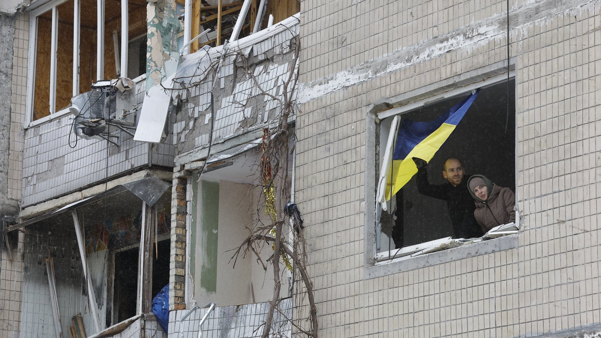 epa11055046 Locals stand by a window with a national flag as they look out from their damaged flat at the site of a damaged building a day after a missile strike, in Kyiv (Kiev), Ukraine, 03 January 2024, amid the Russian invasion. At least 27 people were hospitalized and an injured elderly woman died in an ambulance after a fire broke out in a multi-story building as a result of a rocket attack in Kyiv on 02 January 2024, the city mayor Vitali Klitschko wrote on telegram. Some 130 residents were evacuated from the burning building, the State Emergency Service said.  EPA/SERGEY DOLZHENKO