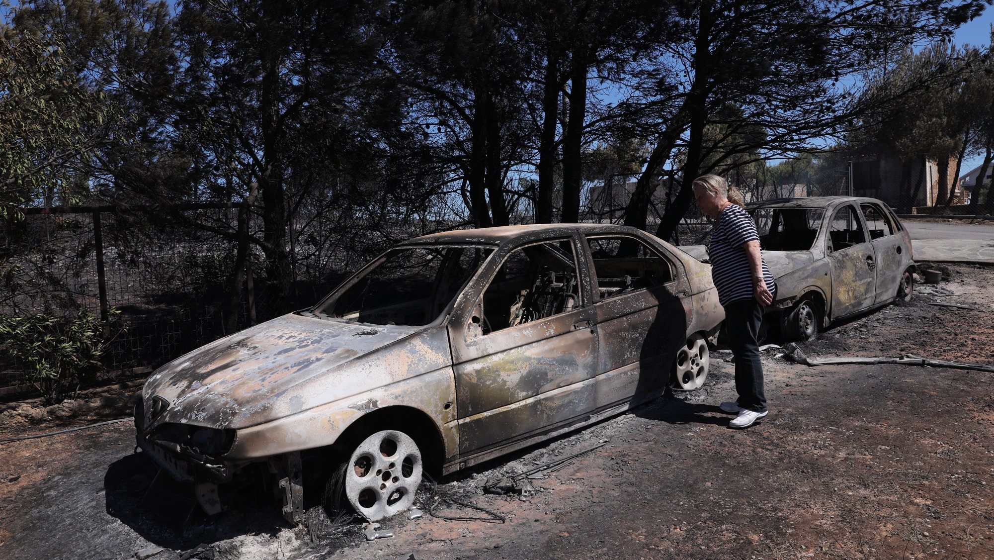 epa10082423 A woman looks at burnt cars after wildfire at the north-east suburb of Anthousa, near Athens, Greece, 20 July 2022. The Penteli fire appears to be subsiding, with no active fronts and all forces now working to extinguish minor small blazes and to prevent any danger of a rekindling of the flames, according to the latest update on 20 July afternoon from the civil protection service&#039;s coordination centre.  EPA/GEORGE VITSARAS