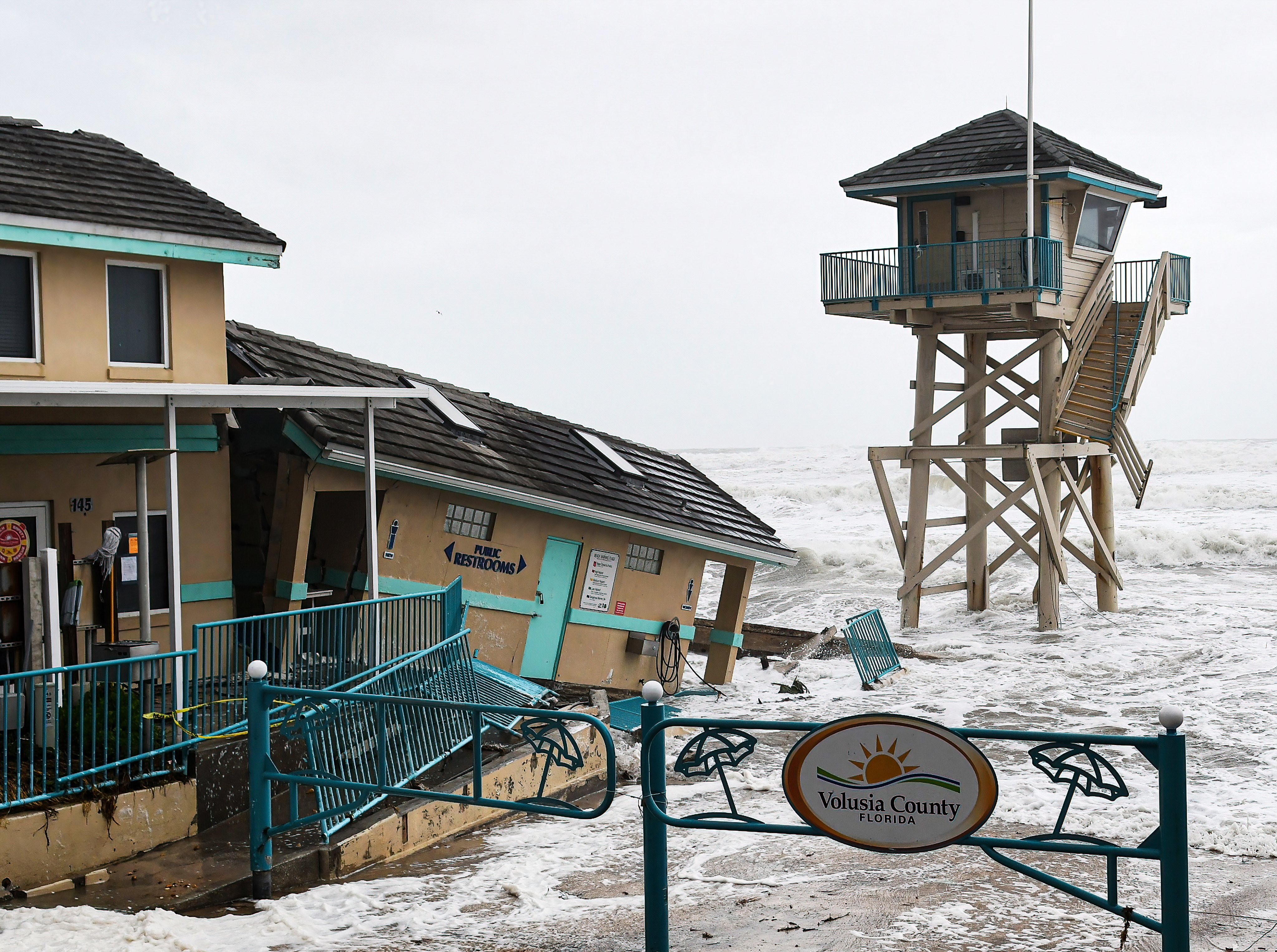 Waves crash near a damaged building and a lifeguard tower in