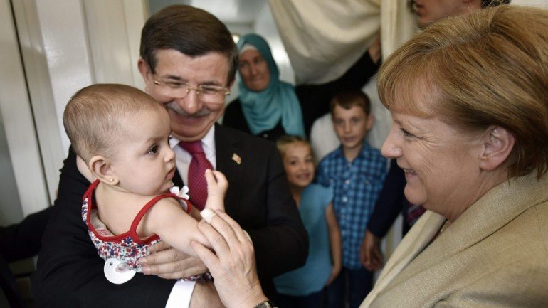 Um bebé sírio é segurado pelo primeiro-ministro da Turquia Ahmet Davutoglu e acariciado por Angela Merkel
