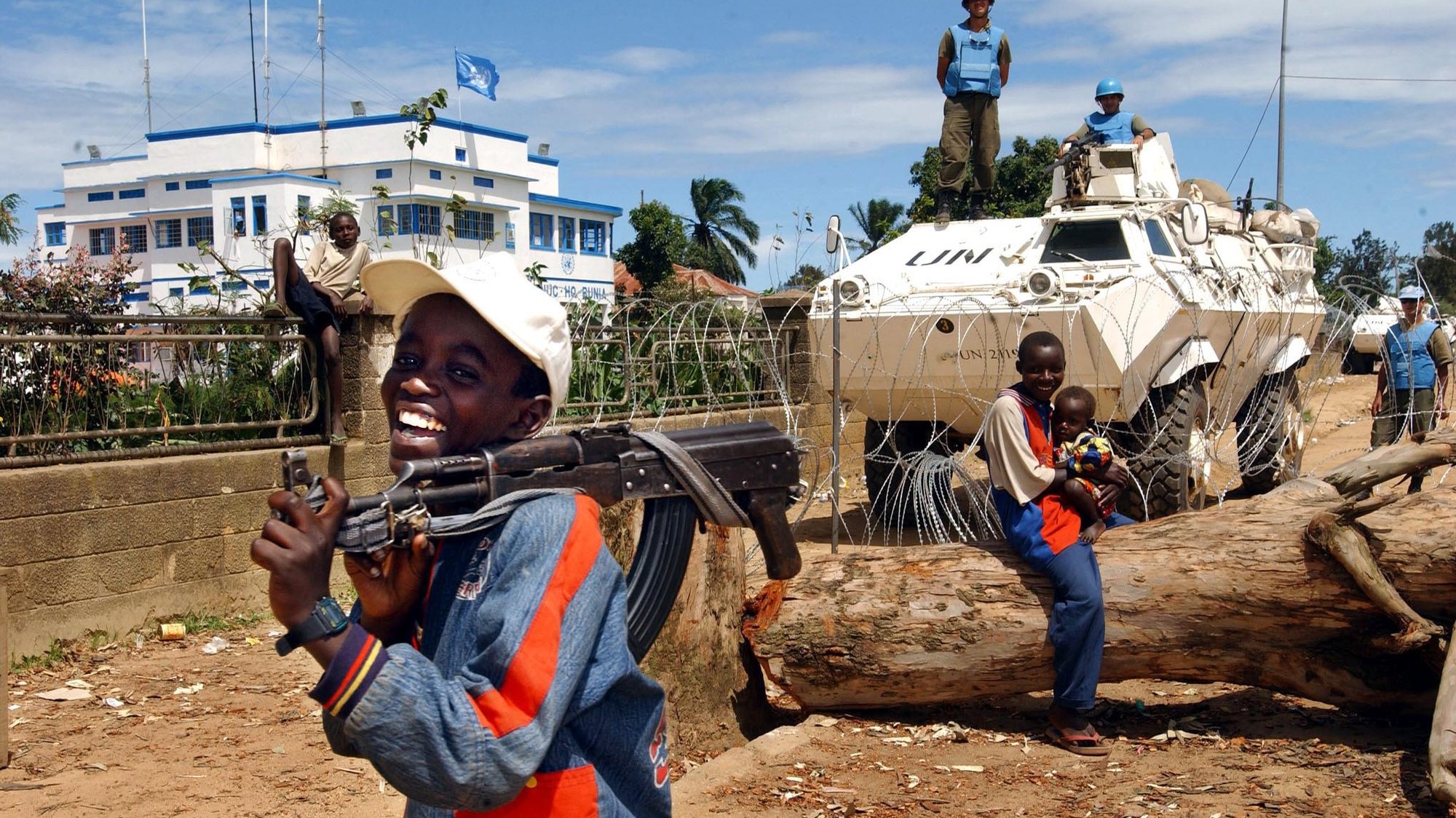 Child soldiers laugh outside the UN compound, 15 May 2003 in Bunia, Democratic Republic of Congo (DRC) as Un soldiers stand guard. The northeastern town of Bunia has been a theater of violent clashes between the Hema and the Lendu militias. MONUC (UN Mission to Congo) compound has been the main assembling point for the displaced from the region.  AFP PHOTO/MARCO L ONGARI             AFP PHOTO/MARCO L ONGARI