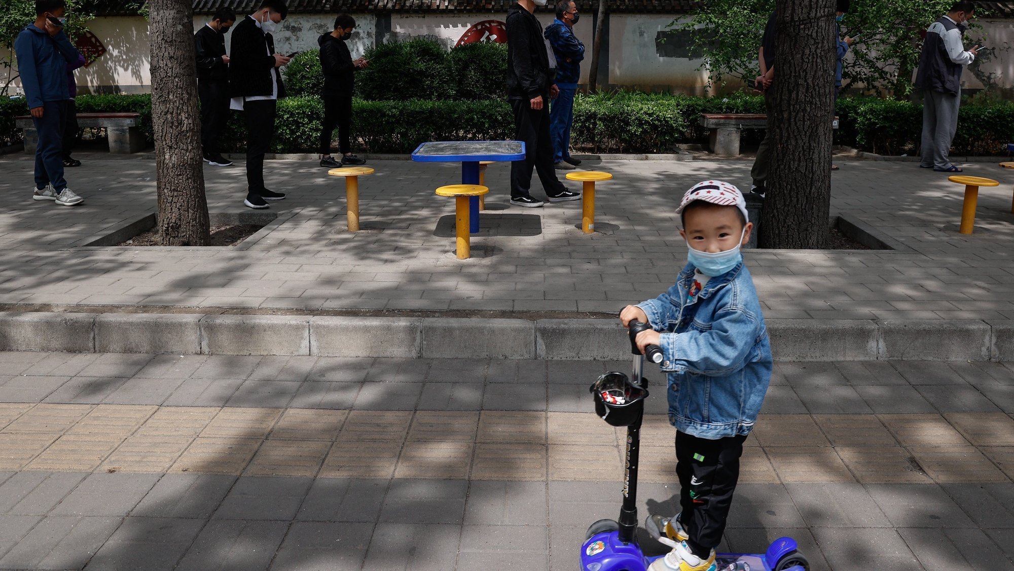 epa09935114 A child rides a scooter along people lining up for a COVID-19 test in Beijing, China, 09 May 2022. Beijing continues its mass testing in an attempt to curb the spread of COVID-19 as dozens of subway stations and bus routes in Beijing remain shut down with public places requiring a negative test result before entering.  EPA/MARK R. CRISTINO