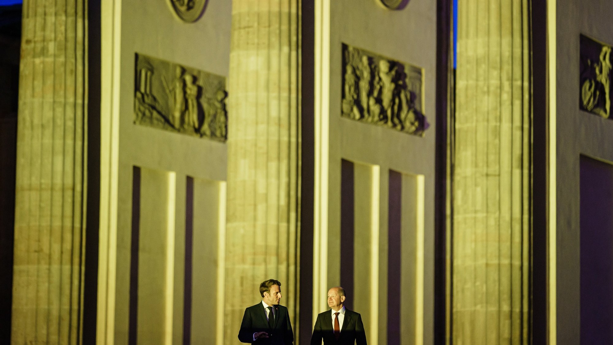 epa09936867 German Chancellor Olaf Scholz (R) and French President Emmanuel Macron (L) talk as they arrive for a photo opportunity in front of the Brandenburg Gate, illuminated in the national colors of Ukraine in Berlin, Germany, 09 May 2022. German Chancellor Olaf Scholz received French President Emmanuel Macron earlier at the Chancellery, where they announced, various buildings all over Europe will be illuminated in the Ukraine colors on Monday evening, among them, the Eiffel tower in Paris.  EPA/CLEMENS BILAN