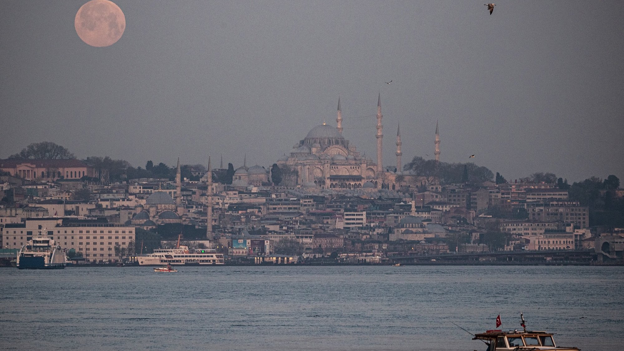 epa09162784 A fisherman labors at the Bosphorus in view of the waning super full moon, next to the Suleymaniye Mosque (C) during sunrise, in Istanbul, Turkey, 27 April 2021. The super full moon is named this way because it is at its closest to earth and thus appears bigger than a normal full moon.  EPA/SEDAT SUNA