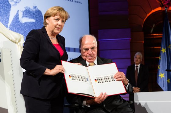BERLIN, GERMANY - SEPTEMBER 27:  German Chancellor Angela Merkel (L) presents a commemorative postal stamp showing former German Chancellor Helmut Kohl as the former Chancellor (R) looks on at a gala evening in Kohl's honour at the Deutsches Museum on September 27, 2012 in Berlin, Germany. Guests from politics, church and society attended the event to honour Kohl on the 30th anniversary of Kohl becoming chancellor. During his chancellorship Kohl facillitated the end of the Cold War, the fall of the Berlin Wall and German reunification.  (Photo by Christian Marquardt - Pool / Getty Images)